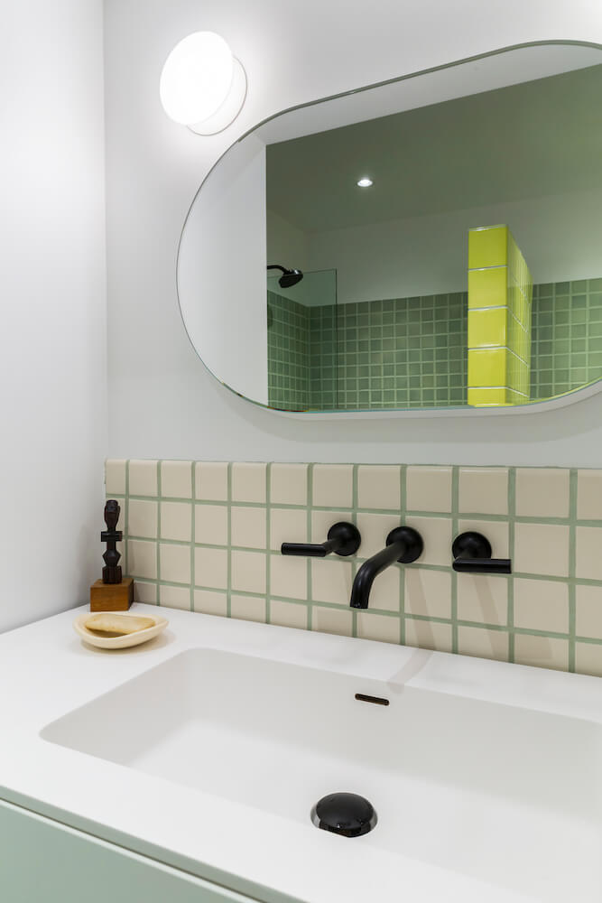 Large white sink with black faucet on cream and green wall tiles along with oval bathroom mirror after renovation