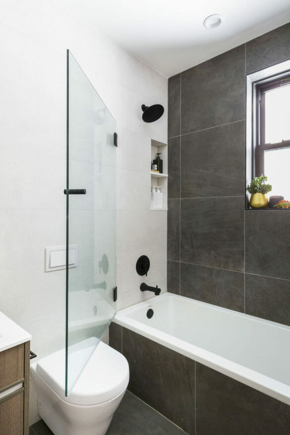 bathroom with dark gray tiles on floor and shower wall and bathtub with black fixtures and glass door and recessed shelves and window after renovation