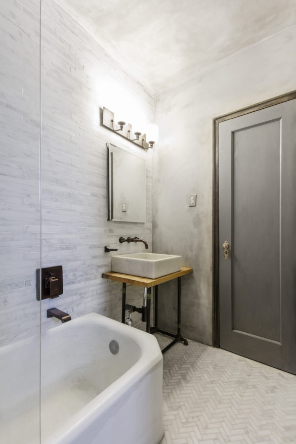 bathroom with light gray tiles on walls and console sink and medicine cabinet with mirror and dark gray door and bathtub with glass wall and herringbone floor tiles after renovation