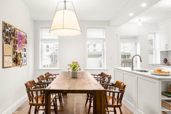 natural brown dining table with chairs in a full white kitchen with white kitchen cabinets and two long windows after renovation